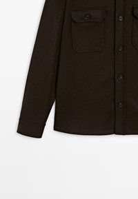 Donkerbruine button-up blouse met lange mouwen, twee borstzakken met knoopflappen, en een eenvoudige manchet op de zichtbare mouw.
