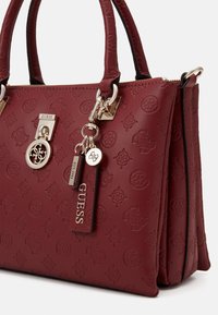 Punainen synteettinen käsilaukku, jossa on kohokuvioitu logokuvio, hopeiset yksityiskohdat, kaksinkertaiset ylhäät kahvat ja vetoketjukiinnitys. Sisältää avaimenperäkoristeet.