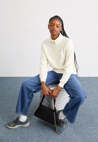 Cream fleece pullover with a half-zip and green accent, paired with relaxed blue denim jeans and multicolored sneakers. Black triangular bag.