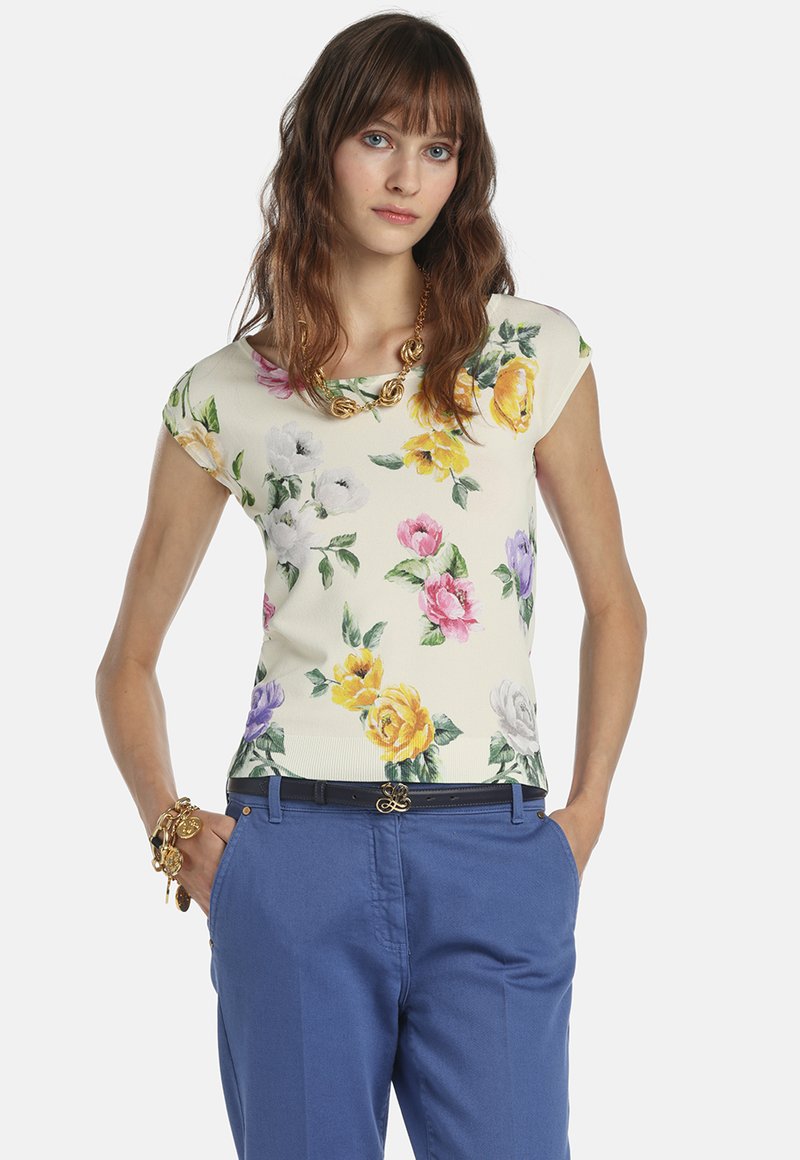 Frau trägt ein cremefarbenes, florales Kurzarmoberteil mit gelben, rosa, weißen und violetten Blumen, blaue Hosen und goldenen Schmuck, die Hände in den Taschen.