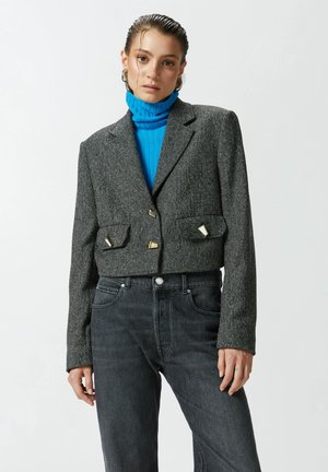 Blazer de tweed gris cortado con botones triangulares dorados, dos bolsillos frontales, combinado con jeans de mezclilla oscura y un jersey de cuello alto azul con canalé.