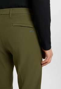 Olivgröna byxor med en slät yta, med en bakficka och en subtil "BOSS"-logotyp i blått, bärs med en svart långärmad topp.