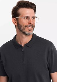 Tmavě šedé polo tričko s zipovým límcem, krátkým rukávem a hladkým povrchem. Na sobě má velké, průhledné brýle a upravený vous.