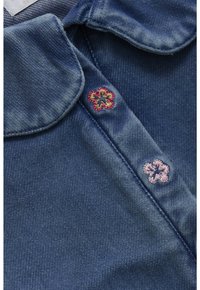 Niebieska tkanina dżinsowa z dwoma małymi haftowanymi kwiatowymi wzorami w pobliżu złożonych brzegów kołnierza, jeden różowy i jeden brązowy z czerwonym konturem.