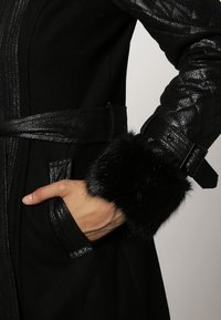 Zwarte jas met een slank ontwerp en een gemêleerde afwerking, bontaccenten bij de manchetten en een ceintuur in de taille, die de gestructureerde silhouet benadrukt.