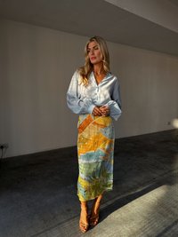 A woman with long, wavy blonde hair stands in a sunlit room, wearing a light blue blouse and a colorful tropical-print skirt, paired with heels.