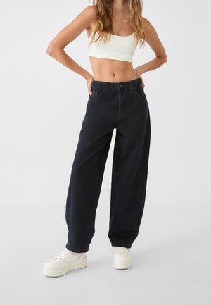 Mujer con vaqueros negros de tiro alto y holgados, top corto blanco y zapatillas blancas con plataforma, de pie con las manos en las caderas frente a un fondo liso.