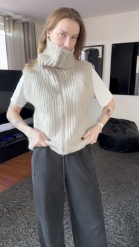 Hellgrauer, ärmelloser Strickpullover mit hohem Kragen, gerippter Textur und dezentem Zopfmuster. Kombiniert mit locker sitzenden schwarzen Hosen.