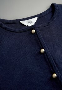 Tmavomodrý pletený kardigán s okrúhlym výstrihom, ozdobený piatimi zlatými gombíkmi na prednej strane a rebrovanou textúrou pozdĺž okrajov. Na štítku je nápis "THE SET."