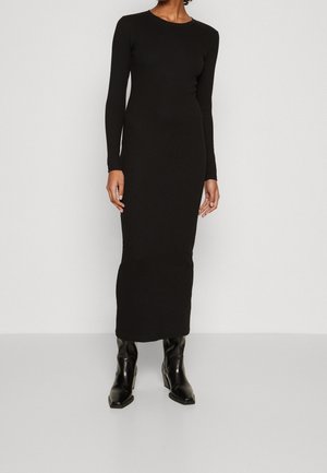 Vrouw draagt een nauwsluitende, lange, enkelhoge zwarte jurk gecombineerd met zwarte laarzen met hak, staand tegen een effen achtergrond.