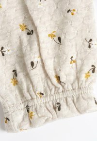 Tissu beige clair avec un motif texturé, présentant de petites fleurs jaunes et blanches ainsi que des feuilles vert foncé. Détail de l'ourlet froncé élastiqué.