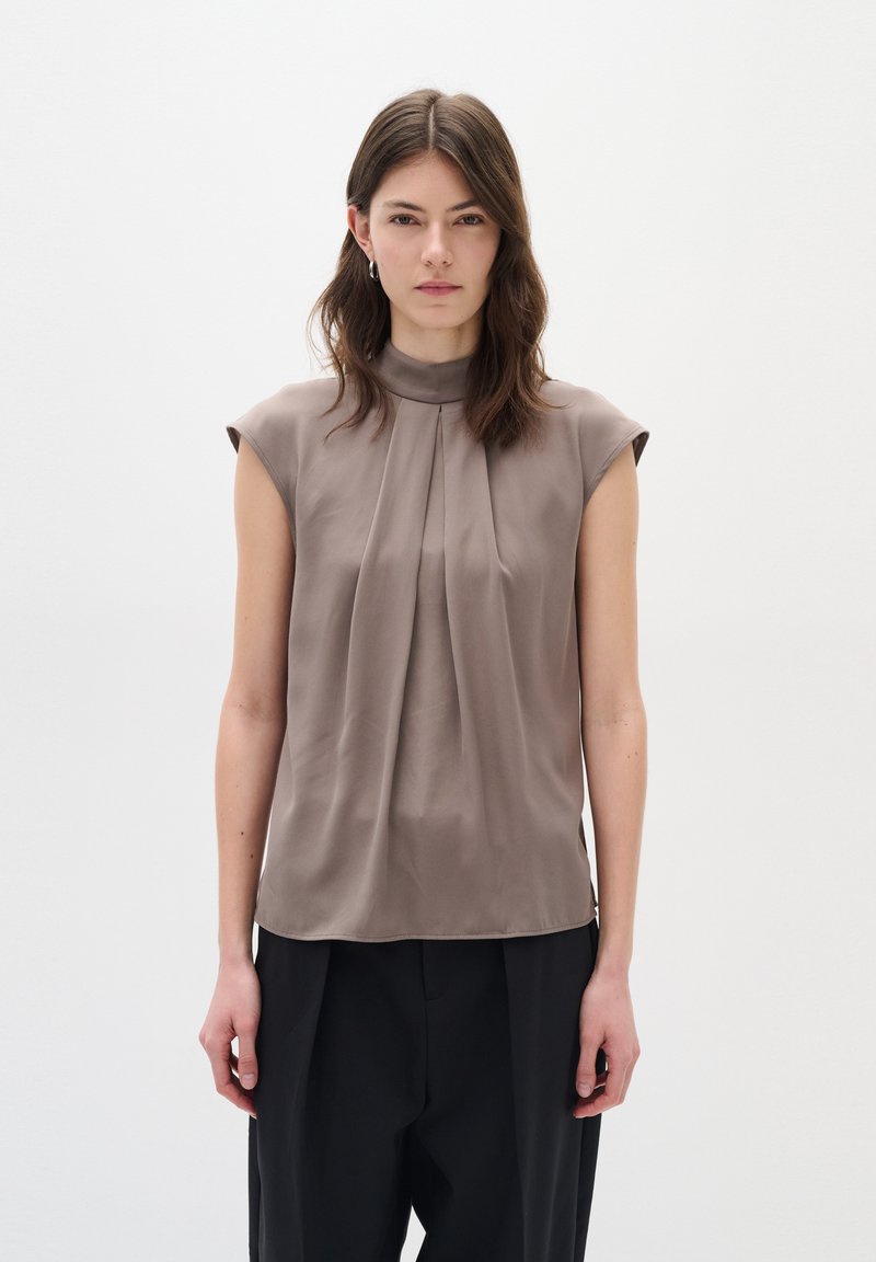 Top senza maniche di colore taupe con scollatura alta e pieghe frontali. Realizzato in tessuto liscio, presenta vestibilità comoda e un design minimalista.