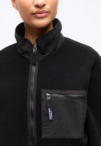 Κοντινό πλάνο ατόμου που φοράει μαύρο φλις μπουφάν με ψηλό γιακά και τσέπη στο στήθος με φερμουάρ, που διαθέτει μαύρη ταινία φερμουάρ με το λογότυπο της μάρκας.