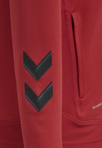 Roter Pullover mit strukturiertem Stoff, einer Reißverschlusstasche und zwei schwarzen Chevron-Akzenten am Ärmel.