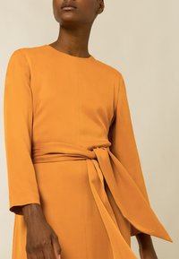 Vestido de manga larga en una cálida tela naranja, con un cuerpo ajustado y un detalle de cintura atada, de textura suave y diseño minimalista.
