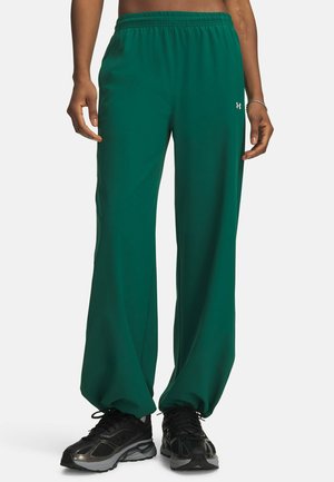 Persoon draagt groene losse broek en zwarte sportschoenen, staand tegen een eenvoudige lichte achtergrond.