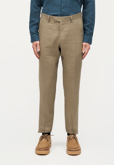 Άνδρας που φοράει μπεζ παντελόνι ίσιας γραμμής, σκούρο μπλε πουκάμισο με μακριά μανίκια, μαύρες κάλτσες και ταμπά σουέντ παπούτσια με κορδόνια, στέκεται με τα χέρια στα πλευρά.