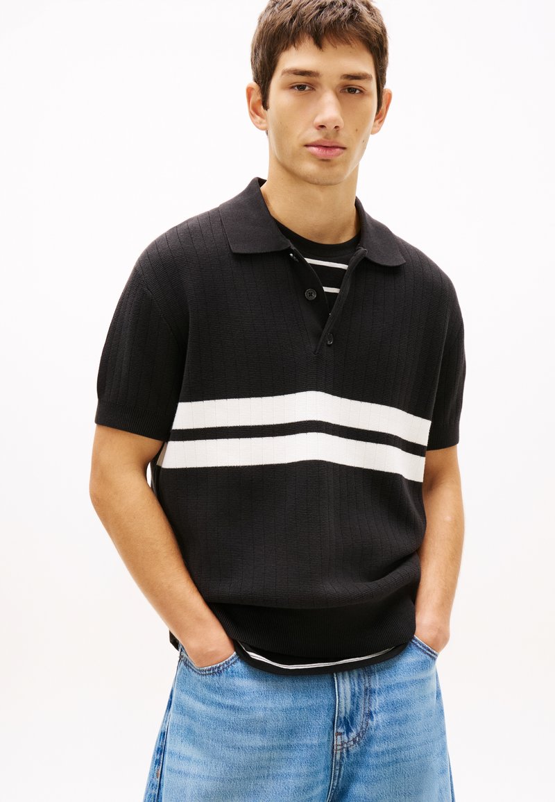 Νεαρός άνδρας με κοντά καστανά μαλλιά, φοράει μαύρο πόλο με λευκές ρίγες και ριγέ πουκάμισο από μέσα, καθώς και ανοιχτόχρωμο τζιν, με τα χέρια στις τσέπες.