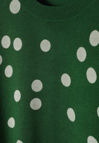 Zelený svetr z texturované tkaniny, zdobený rozptýlenými bílými puntíky a žebrovaným límcem. Detailní pohled zvýrazňuje vzor.