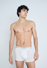 Boxer briefs de algodón blanco con una cinturilla suave que presenta el texto "Calvin Klein". El modelo está sin camiseta y mirando ligeramente de lado.