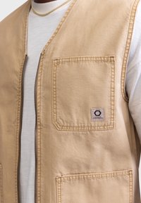 Chaleco de cremallera en color tostado, con un bolsillo frontal cuadrado, acentos cosidos y un parche de logo. Fabricado con un tejido duradero, presenta un ajuste estándar.