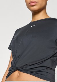 Μαύρο αθλητικό μπλουζάκι με μπροστινό κόμπο, κατασκευασμένο από λείο ύφασμα, με μικρό λευκό λογότυπο Nike στο στήθος.
