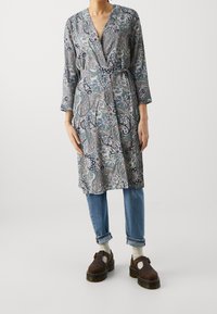 Długa, wzorzysta sukienka kopertowa w odcieniach niebieskiego i taupe z wzorem paisley, z dekoltem w serek, długimi rękawami i wiązaniem w talii, zestawiona z dżinsami.