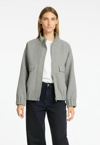 Grijs zip-up jack met een opstaande kraag, textuurstof, twee voorzakken en gathered mouwen, gecombineerd met donkere broek en een wit shirt eronder.