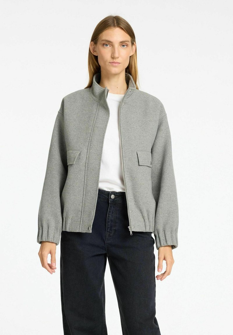 Grijs zip-up jack met een opstaande kraag, textuurstof, twee voorzakken en gathered mouwen, gecombineerd met donkere broek en een wit shirt eronder.