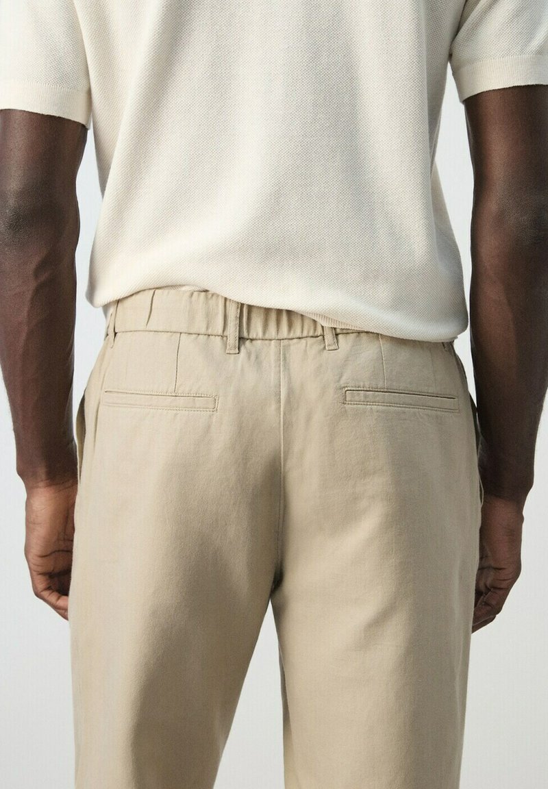 Donkere huidskleur, gekleed in een wit gebreid T-shirt met korte mouwen, ingestopt in beige op maat gemaakte broek met achterzakken met lijst.