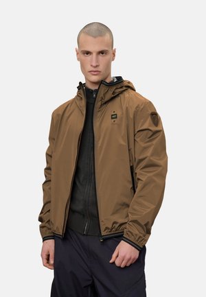 Giovane uomo che indossa una giacca marrone con cappuccio sopra un indumento nero con cerniera e pantaloni scuri, in piedi davanti a uno sfondo uniforme.