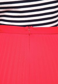 La falda roja plisada presenta una textura suave y un cierre de cremallera en la parte posterior, combinada con una blusa a rayas azul marino y blanca.
