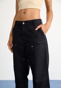 Pantalones de pierna ancha negros de algodón, con bolsillos frontales, herrajes plateados y detalles de costura. Textura suave con un brillo sutil.