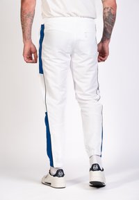 Pantalons de sport blancs avec des panneaux latéraux bleus, une taille élastique et un passepoil noir. Le modèle se tient avec une chaussure neutre, montrant la vue de dos.