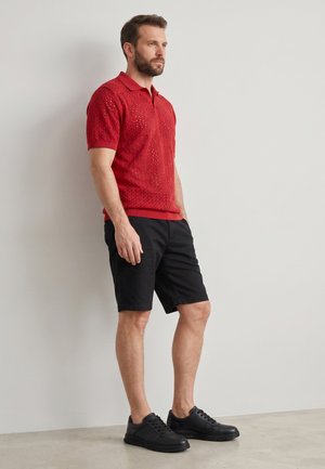 Mann trägt ein rotes kurzärmeliges Strick-Polo, schwarze Shorts und schwarze Sneakers, steht vor einer schlichten weißen Wand.