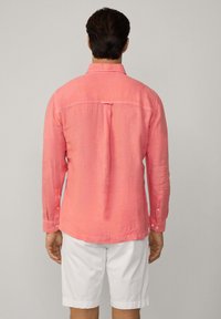 Camicia lunga manica in lino rosa con polsini abbottonati e orlo arrotondato, dotata di una piega centrale sul retro e colletto. Indossata con pantaloni corti bianchi.