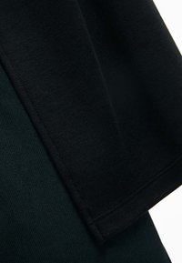 Svart tyg hänger över mörkgrönt tyg och har en slät yta med en synlig söm. Materialen verkar mjuka och tjocka.