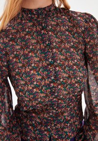 Langærmet blouse med høj krave, prydet med et mørkt blomstermønster i orange, pink og grønt på en sort baggrund. Dekorative knapper inkluderet.