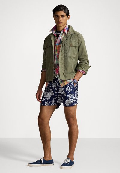Veste utilitaire verte, chemise à boutons à carreaux, short à motifs floraux navy, et baskets navy. Les caractéristiques du style incluent des poches et des motifs colorés.