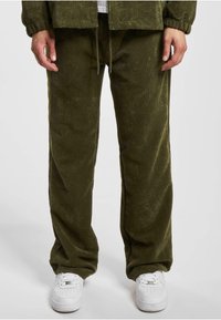 Pantalones de corduroy verde oliva con textura de ribete vertical; cintura con cordón; corte relajado; combinados con zapatillas blancas para crear contraste.