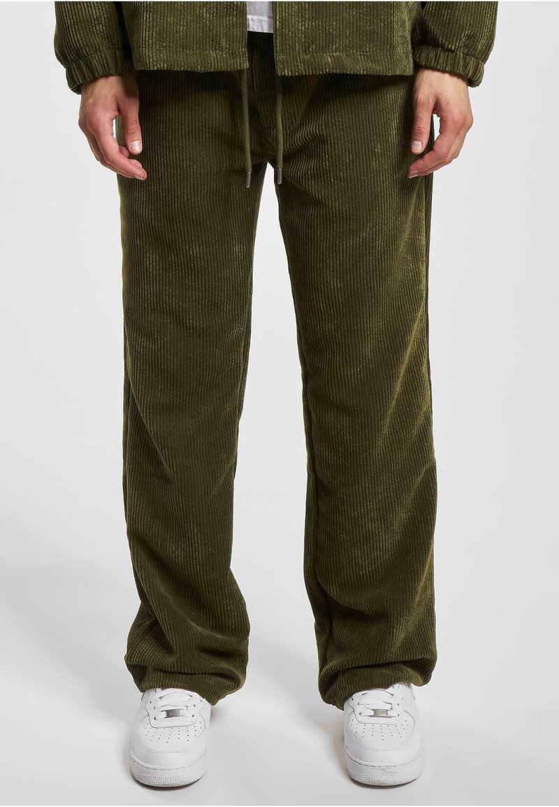 Pantalones de corduroy verde oliva con textura de ribete vertical; cintura con cordón; corte relajado; combinados con zapatillas blancas para crear contraste.