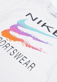 Balta kokvilnas t-krekls ar gradienta logo, kas sastāvno zilajiem, purpura un oranžiem toņiem, papildināts ar uzsvērtiem melniem burtiem, kas lasāms "NIKE SPORTSWEAR."