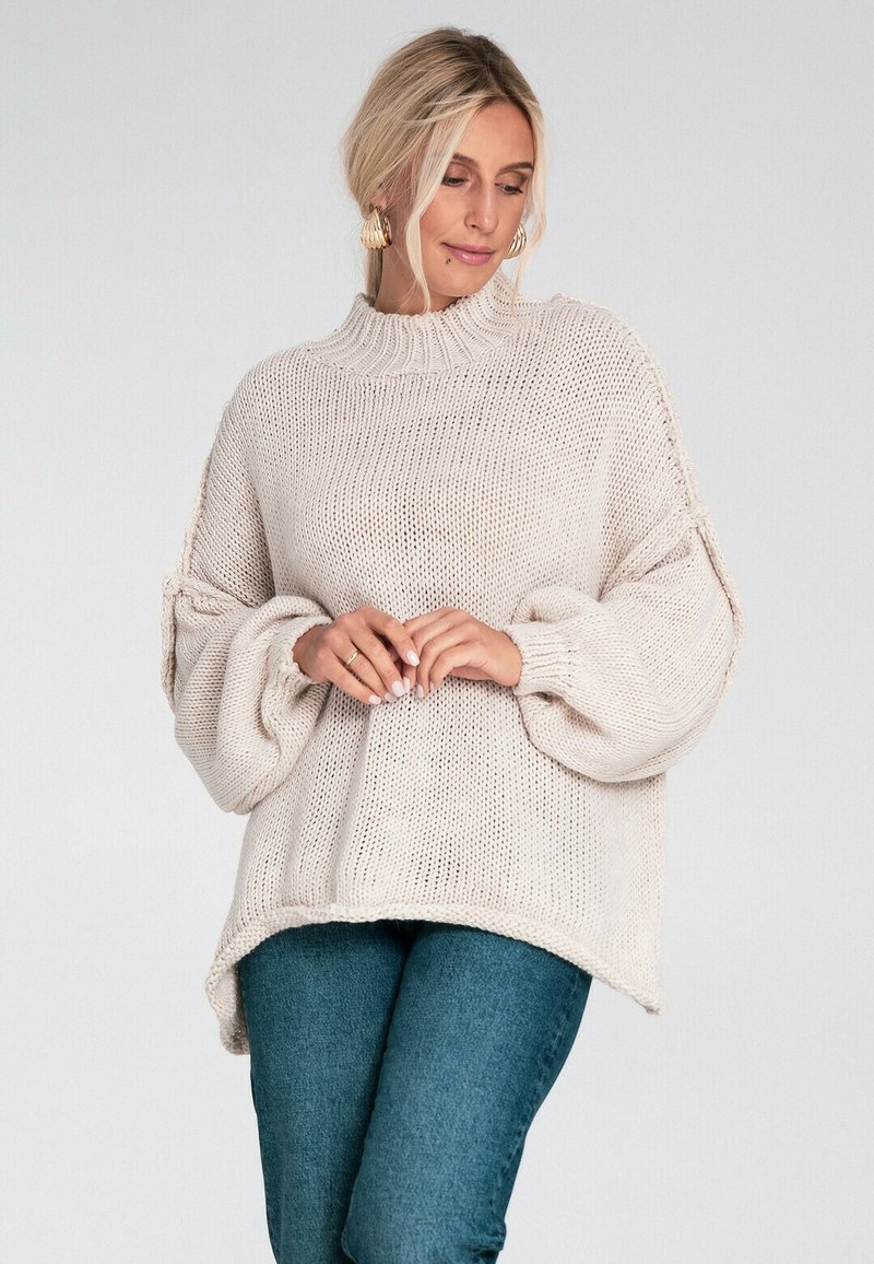 Jasno beżowy sweter z dzianiny o wysokim kołnierzu, z oversize'owymi rękawami w kształcie balonu oraz luźnym krojem. Posiada teksturowany, otwarty splot.
