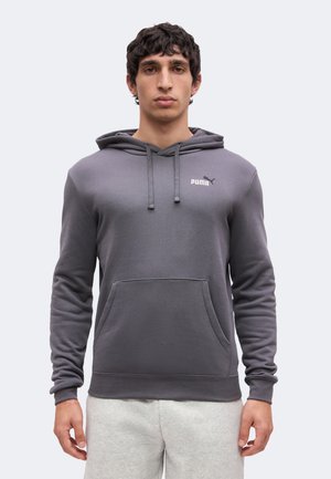 Jonge man draagt een donkergrijze Puma hoodie met voorzak en lichtgrijze sweatbroek, staand tegen een effen lichte achtergrond.