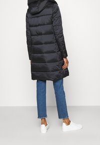 Schwarzer Steppmantel mit Kapuze, mit gestepptem Design und mittellangem Schnitt, kombiniert mit blauen Jeans und weißen Turnschuhen.