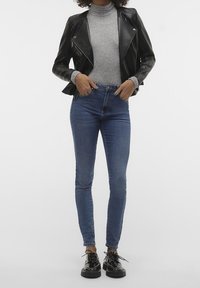 Zwarte leren motorjas, grijze nauwsluitende coltrui en blauwe strakke spijkerbroek. Persoon draagt glanzende zwarte veterschoenen. Eenvoudige achtergrond.