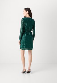 Abito verde con paillettes, maniche lunghe, scollo rotondo, vita elastica e orlo dritto. Indossato con sandali neri con tacco alto.