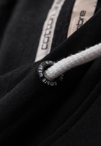 Černá bavlněná látka s bílým stahovacím šňůrou, která má kovový průchod s vyrytým nápisem "wear". Švy jsou viditelné podél okrajů.