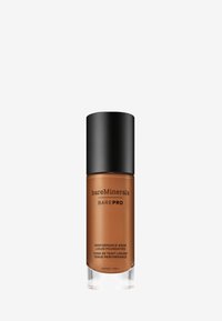 Fondotinta liquido in una bottiglia cilindrica trasparente con tappo a vite nero. L'etichetta presenta il nome del marchio e la descrizione del prodotto in testo nero.