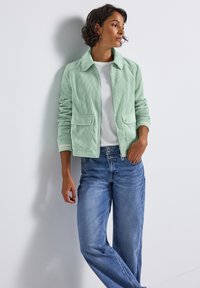 Hellgrüne Cordjacke mit Kragen, zwei Fronttaschen und Reißverschluss, getragen über einem weißen Hemd und kombiniert mit blauen Jeans.